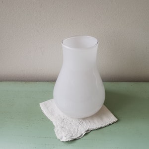 May include: A white frosted glass vase with a rounded bottom and a narrow neck. The vase is sitting on a white doily on a green surface.