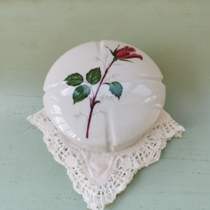 May include: White ceramic trinket box with a single red rose and green leaves design. The box is sitting on a white lace doily.