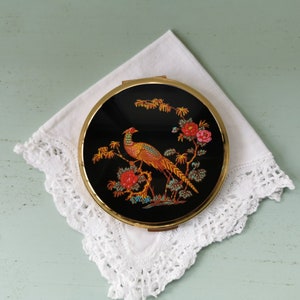 Può includere: Uno specchio compatto dorato con sfondo nero e un'immagine colorata di un fagiano e fiori.