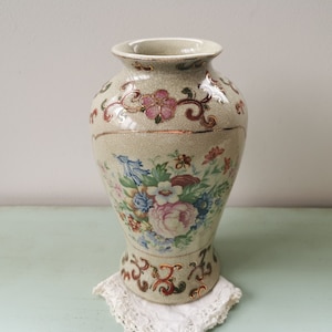 May include: A cream-colored ceramic vase with a floral pattern in shades of pink, red, blue, and green. The vase has a textured surface and gold accents.