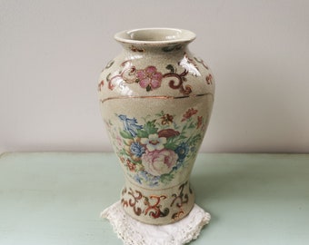 May include: A cream-colored ceramic vase with a floral pattern in shades of pink, red, blue, and green. The vase has a textured surface and gold accents.
