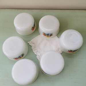 May include: Six white ceramic ramekins with floral designs. The ramekins are arranged in a circle on a light green surface.