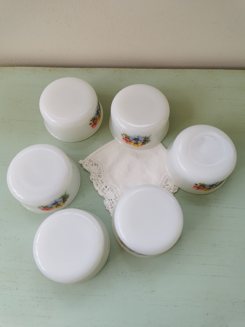 May include: Six white ceramic ramekins with floral designs. The ramekins are arranged in a circle on a light green surface.