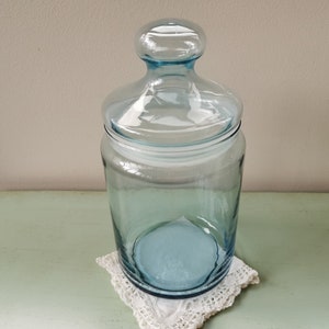 Puede incluir: Un tarro de vidrio azul claro con tapa. El tarro está sobre un encaje de ganchillo blanco.