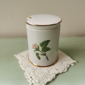 May include: White ceramic jar with a gold trim and a pink rose design. The jar has a lid with a small spout.