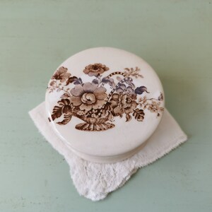 Peut inclure: Un couvercle en céramique blanche avec un motif floral brun représentant un panier de fleurs. Le couvercle est posé sur une dentelle blanche.