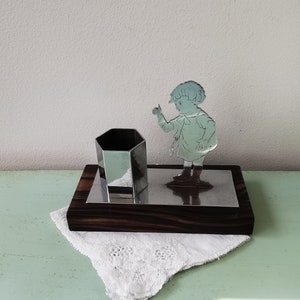 Puede incluir: Un organizador de escritorio de plata con un porta lápices hexagonal y un recorte de un niño apuntando al porta lápices. El organizador está sobre una base de madera oscura.