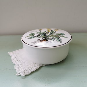 May include: White ceramic round trinket box with a floral design on the lid. The lid features a green plant with yellow flowers and brown roots. The box is sitting on a white crocheted doily.
