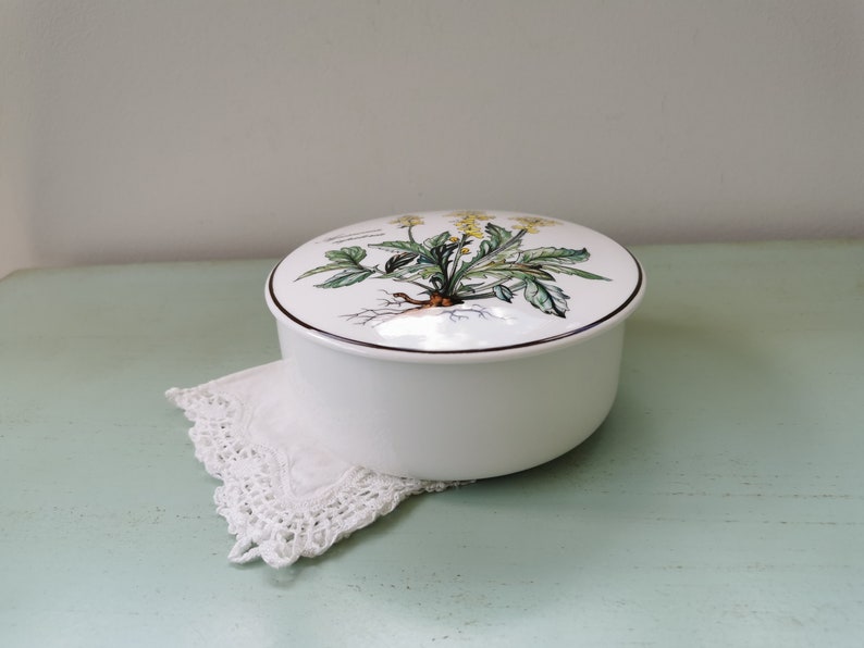May include: White ceramic round trinket box with a floral design on the lid. The lid features a green plant with yellow flowers and brown roots. The box is sitting on a white crocheted doily.
