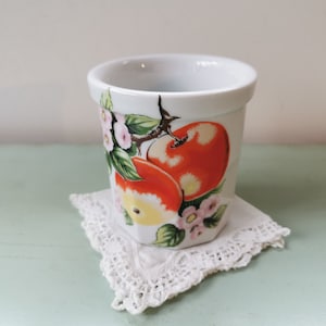 Peut inclure: Une tasse en céramique blanche avec un motif de pommes rouges et jaunes. La tasse a une tige et des feuilles vertes et des fleurs roses.