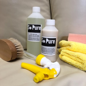May include: Two white bottles of So Pure Leather Cleaner, a yellow spray bottle, a wooden brush, and yellow and peach microfiber cloths are arranged on a beige surface. The bottles have black and white labels. The spray bottle is yellow and white.