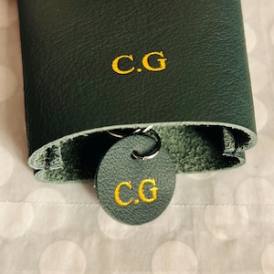 May include: Dark green leather key holder with the embroidered initials "C.G" in gold. A matching round key tag with the same initials hangs from a silver key ring. The key holder is folded over, revealing a lighter green interior.