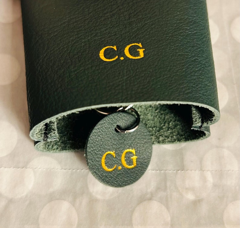 May include: Dark green leather key holder with the embroidered initials "C.G" in gold. A matching round key tag with the same initials hangs from a silver key ring. The key holder is folded over, revealing a lighter green interior.