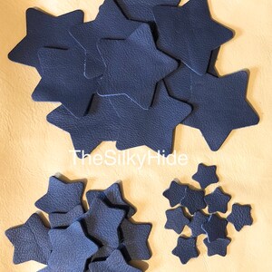 May include: A collection of navy blue leather star shapes in various sizes. The stars are arranged in three groups on a beige surface. The text "TheSilkyHide" is visible in the centre of the image.