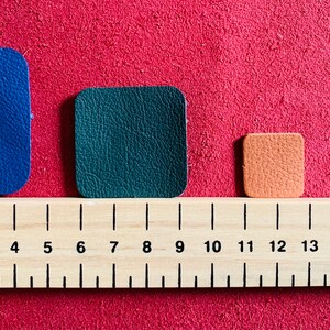 May include: Three leather squares of different sizes and colors: blue, green, and orange. The squares are lined up on a wooden ruler marked in centimeters.