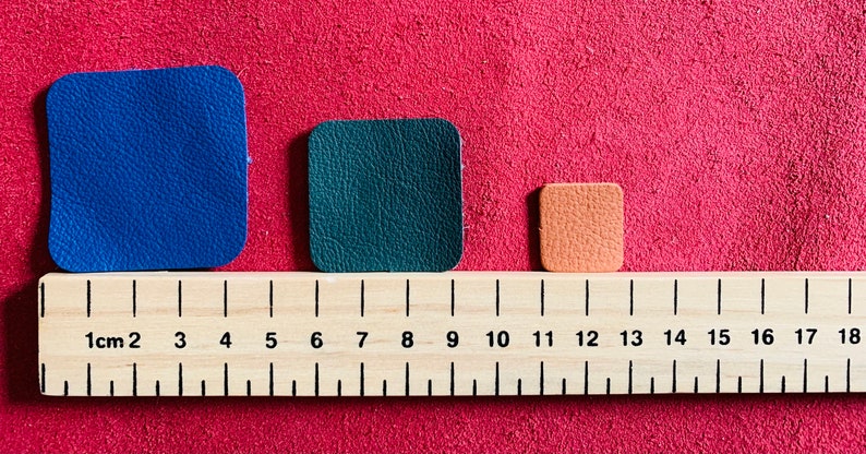 May include: Three leather squares of different sizes and colors: blue, green, and orange. The squares are lined up on a wooden ruler marked in centimeters.