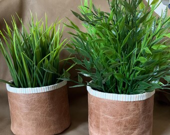 May include: Two brown leather plant pots with white and orange striped fabric trim. The pots are filled with green artificial plants.