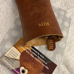 May include: A brown leather key holder with the initials "MJH" embossed on the front. A matching key tag with the same initials is attached. The key holder is open, revealing the interior. A business card is visible.