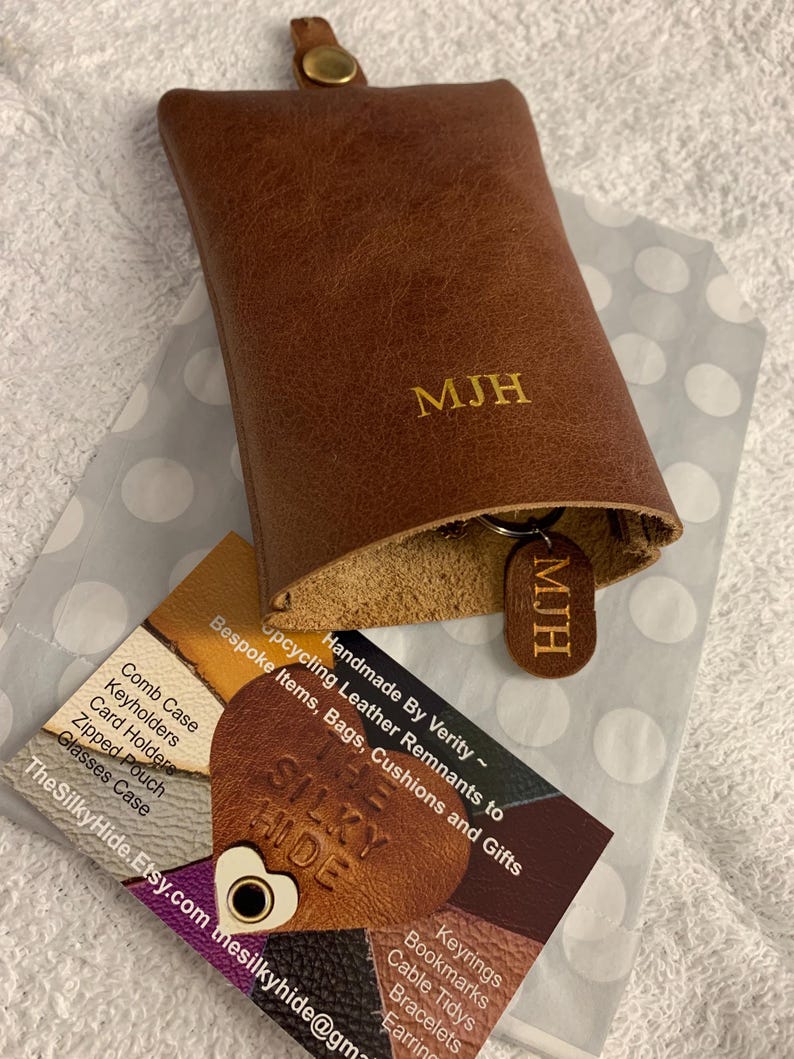 May include: A brown leather key holder with the initials "MJH" embossed on the front. A matching key tag with the same initials is attached. The key holder is open, revealing the interior. A business card is visible.