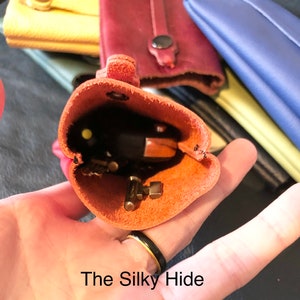 May include: An open, orange leather key holder with keys inside, held in a hand. The text "The Silky Hide" is at the bottom. Other leather items in various colors are in the background.