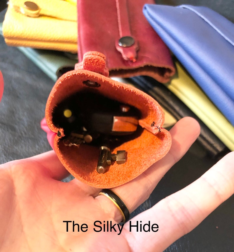 May include: An open, orange leather key holder with keys inside, held in a hand. The text "The Silky Hide" is at the bottom. Other leather items in various colors are in the background.