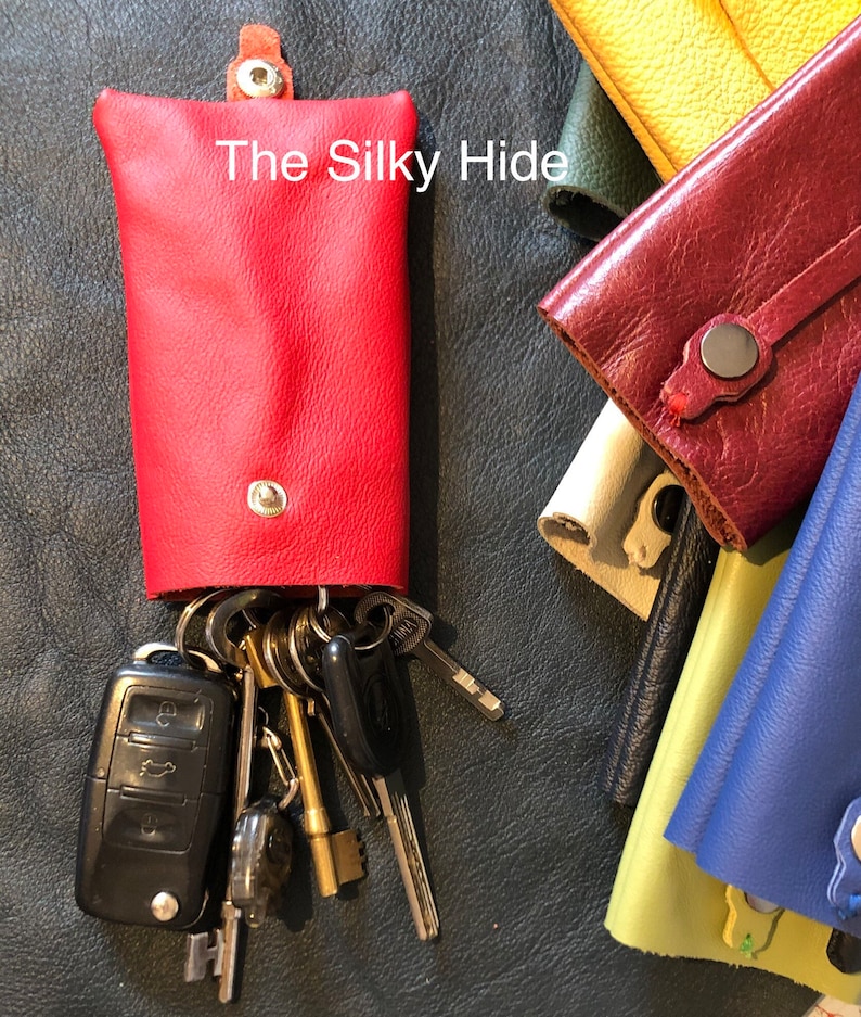 May include: A red leather key holder with a snap closure, holding a set of keys and a car key fob. The text "The Silky Hide" is printed on the front. Other colored key holders are visible in the background.