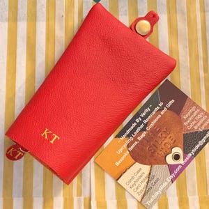 May include: A red leather key holder with the initials "KT" in gold lettering. The key holder has a snap closure and a small tag with the same initials. A business card is next to the key holder.