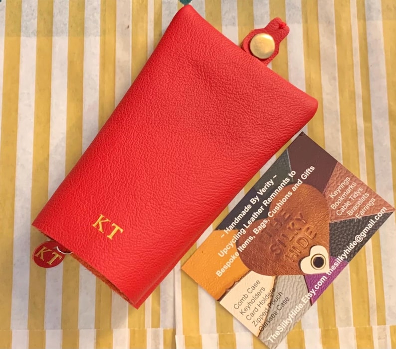 May include: A red leather key holder with the initials "KT" in gold lettering. The key holder has a snap closure and a small tag with the same initials. A business card is next to the key holder.