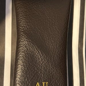 May include: A brown leather glasses case with the initials "AJL" embroidered in gold thread. The case has a textured surface and a curved shape, likely designed to protect eyewear. The background features a black and white striped pattern.