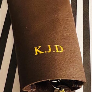 May include: A brown leather key holder with the embroidered initials "K.J.D" in gold. The key holder is closed, with a small leather tag with the same initials visible. The background has black and white stripes.