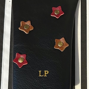 May include: Black leather wallet with four star-shaped embellishments in red and brown, each with a brass-colored center. The initials "LP" are embossed in gold on the front. The wallet is closed.