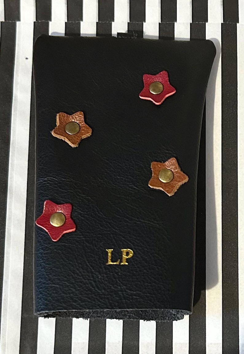 May include: Black leather wallet with four star-shaped embellishments in red and brown, each with a brass-colored center. The initials "LP" are embossed in gold on the front. The wallet is closed.