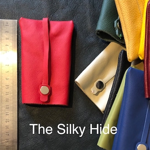 May include: A red leather key holder with a silver snap closure and a strap. The key holder is rectangular and has a smooth texture. Other colorful leather key holders are visible in the background. The text "The Silky Hide" is at the bottom.