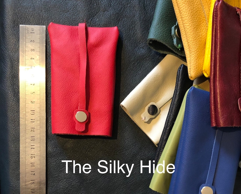 May include: A red leather key holder with a silver snap closure and a strap. The key holder is rectangular and has a smooth texture. Other colorful leather key holders are visible in the background. The text "The Silky Hide" is at the bottom.
