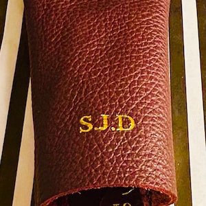 May include: A burgundy leather key holder with the initials "S.J.D" embroidered in gold. The key holder has a textured surface and a loop at the top. The background features a black and white striped pattern.