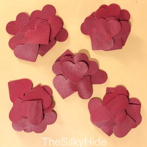 May include: A collection of six piles of burgundy leather heart shapes. The hearts are arranged in a semi-circle on a light beige surface. The text "TheSilkyHide" is visible at the bottom of the image.