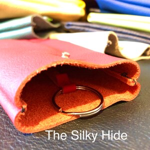 May include: A red leather key holder with a silver key ring peeking out. The key holder is open, revealing the interior. The text "The Silky Hide" is visible at the bottom. Other leather items in various colors are in the background.