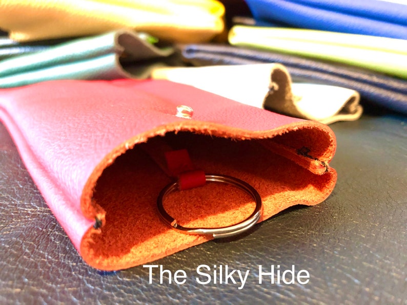 May include: A red leather key holder with a silver key ring peeking out. The key holder is open, revealing the interior. The text "The Silky Hide" is visible at the bottom. Other leather items in various colors are in the background.