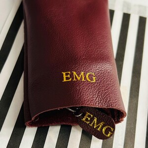 May include: A burgundy leather key holder with gold embroidered initials "EMG". The key holder is rolled up, revealing a matching key fob with the same initials. The background features a black and white striped pattern.