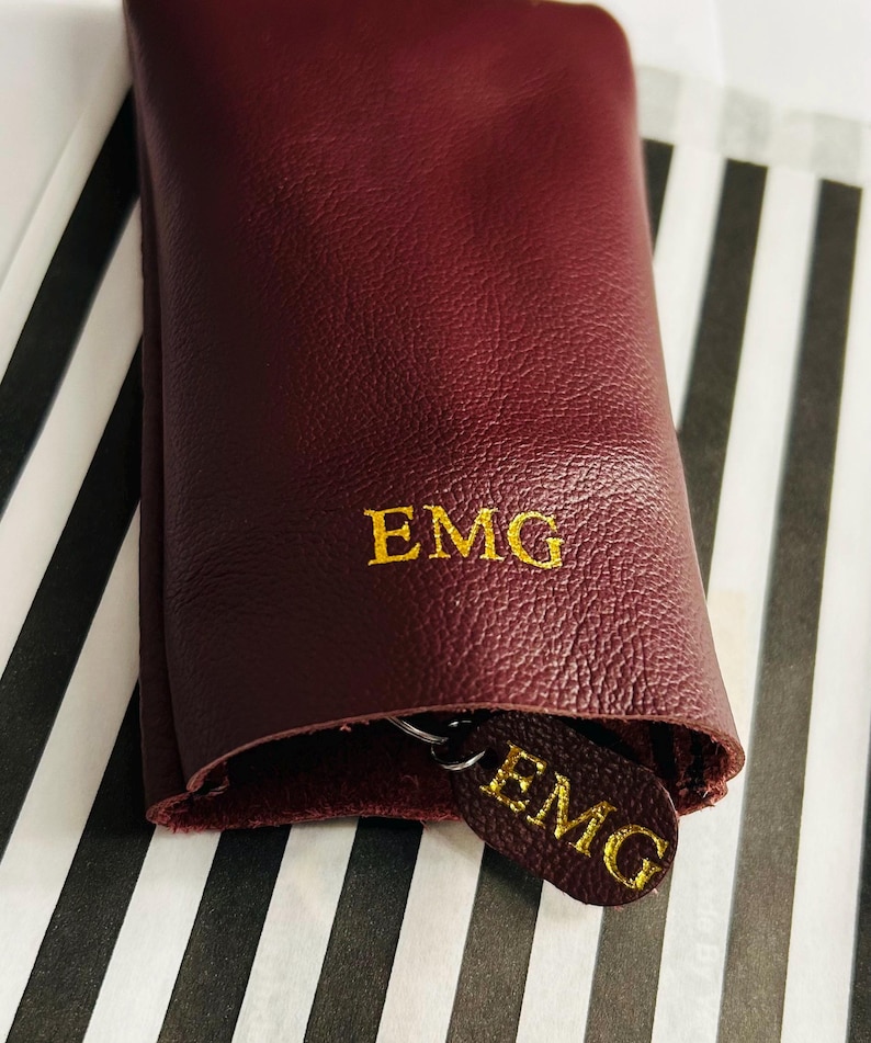 May include: A burgundy leather key holder with gold embroidered initials "EMG". The key holder is rolled up, revealing a matching key fob with the same initials. The background features a black and white striped pattern.