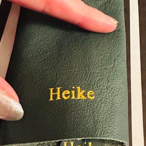 May include: Dark green leather item with the name "Heike" embroidered in gold thread. The item is held by a hand, and the background features a striped pattern.