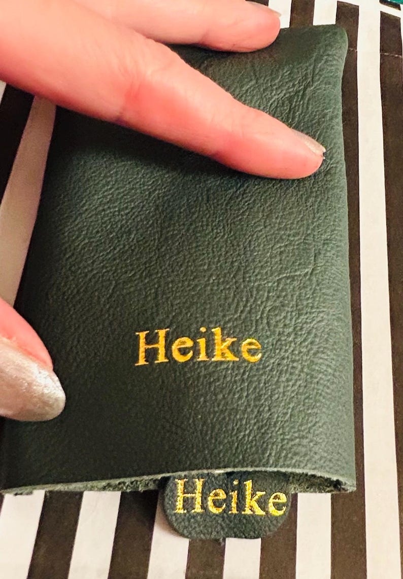 May include: Dark green leather item with the name "Heike" embroidered in gold thread. The item is held by a hand, and the background features a striped pattern.