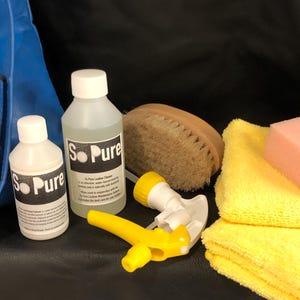 May include: A leather care kit featuring two bottles of "So Pure" leather cleaner, a yellow spray bottle, a brush, a pink sponge, and yellow microfiber cloths. A blue leather bag is in the background. The kit is for leather maintenance.