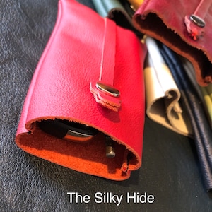 May include: A close-up of a red leather key holder with a silver snap closure. The key holder is open, revealing a key inside. The text "The Silky Hide" is at the bottom of the image. Other leather items are in the background.