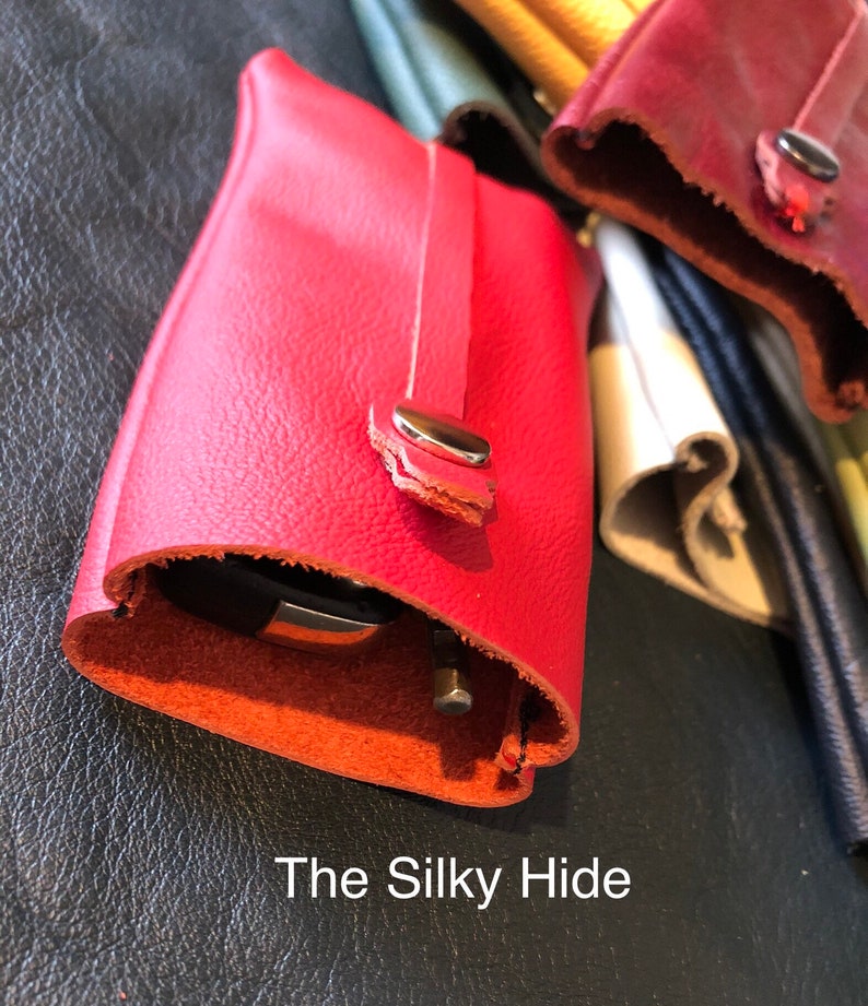 May include: A close-up of a red leather key holder with a silver snap closure. The key holder is open, revealing a key inside. The text "The Silky Hide" is at the bottom of the image. Other leather items are in the background.