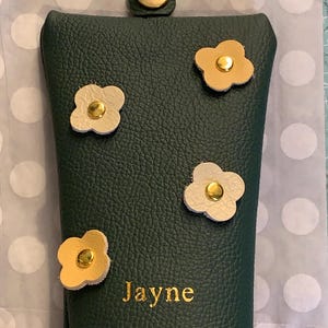 May include: A dark green leather eyeglass case with four flower appliqu&eacute;s in cream and gold. The case has a gold snap closure and the name "Jayne" is embossed in gold lettering.