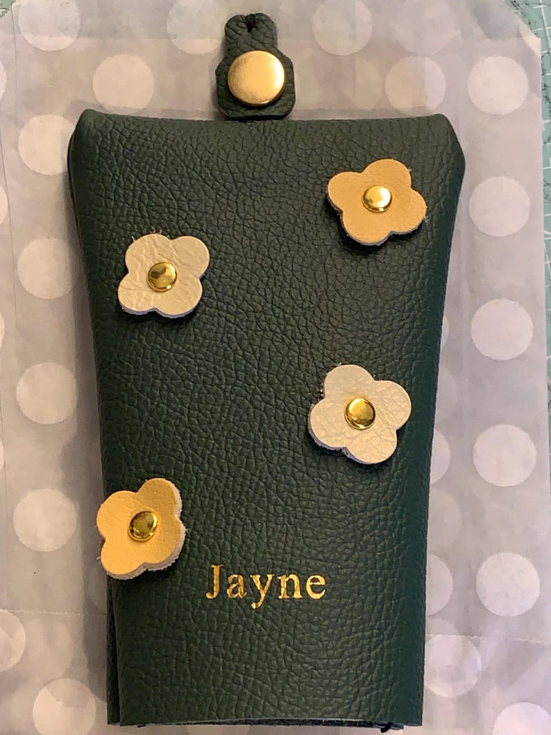 May include: A dark green leather eyeglass case with four flower appliqu&eacute;s in cream and gold. The case has a gold snap closure and the name "Jayne" is embossed in gold lettering.