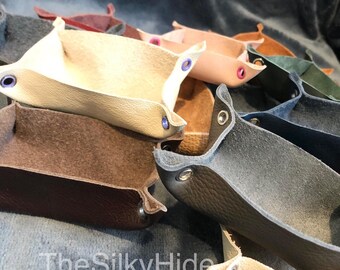 May include: A collection of small, leather trays in various colors, including brown, beige, gray, and black. Each tray has a rounded, rectangular shape and features a small, metal eyelet on each corner. The trays are stacked on top of each other, with the text "The Silky Hide" visible in the bottom right corner.