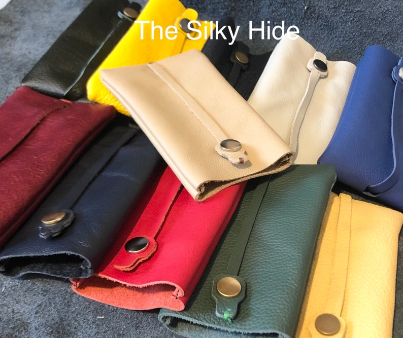 May include: A collection of colorful leather key holders in various shades, including red, yellow, green, and blue. Each holder features a snap closure and a strap detail. The text "The Silky Hide" is visible in the image.