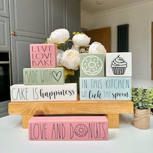 May include: A selection of painted wooden blocks with kitchen-related designs and text. The blocks, in pastel shades, display phrases such as "Live Love Bake" and "Cake is Happiness." The blocks are arranged on a wooden stand.